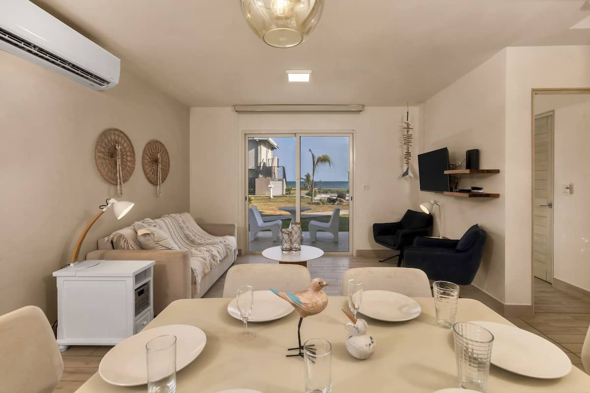 Una sala moderna con comedor, un sofá, un televisor montado en la pared y vista al océano a través de la puerta de vidrio.