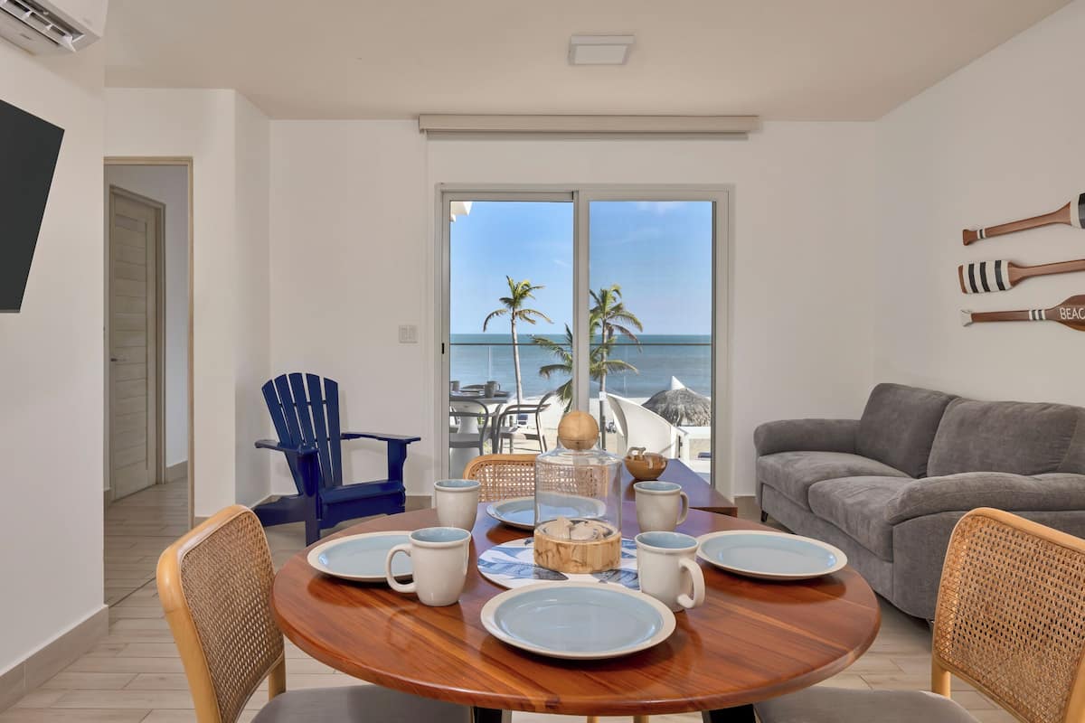 Un comedor con una mesa de madera dispuesta para cuatro, un sofá gris y vista a la playa a través de amplios ventanales correderas.