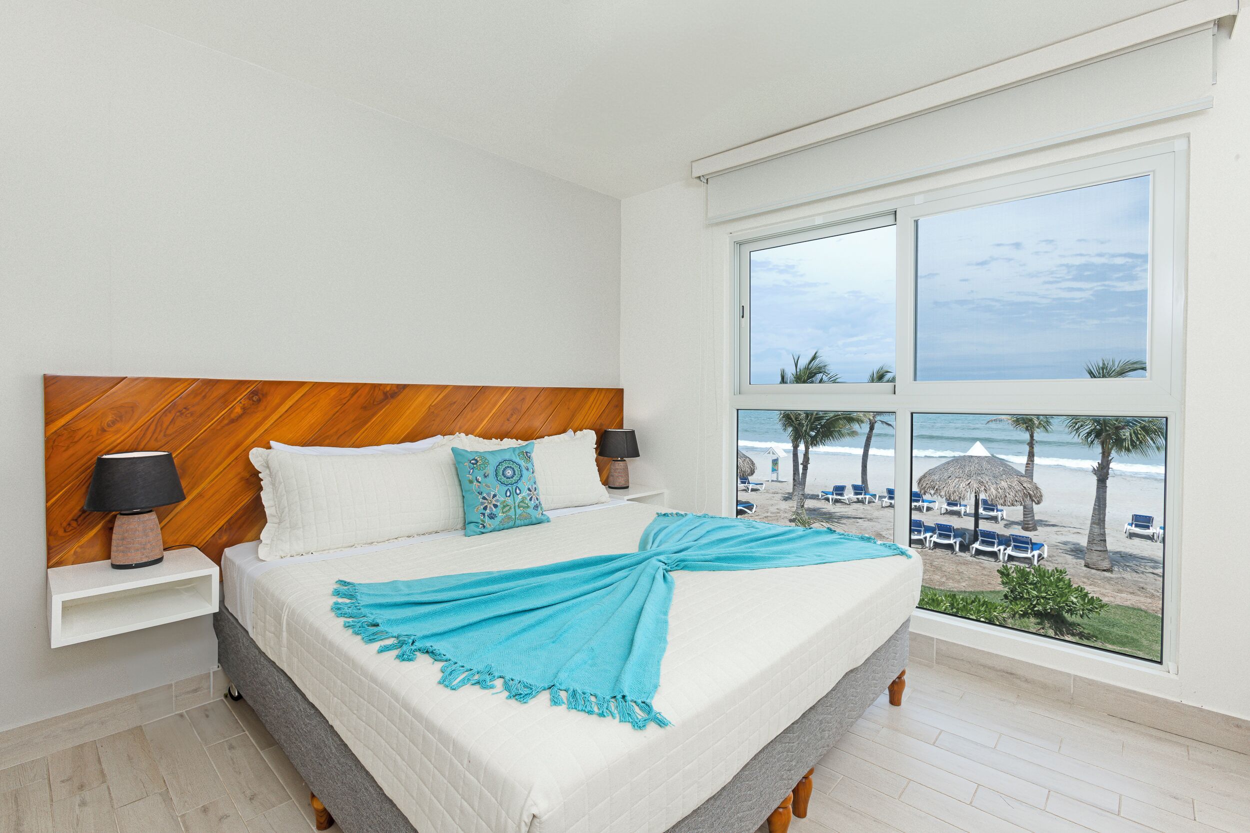 A bedroom with a large bed, a wooden headboard, a nightstand with a lamp, and a view of the beach through large windows.