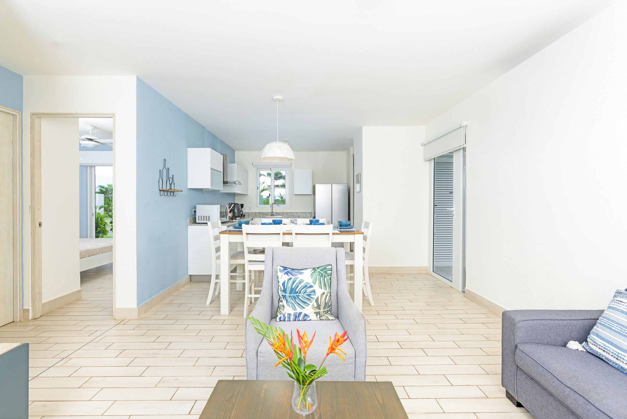 A modern living room with a grey sofa, a dining area with a white table and chairs, and a kitchen in the background.