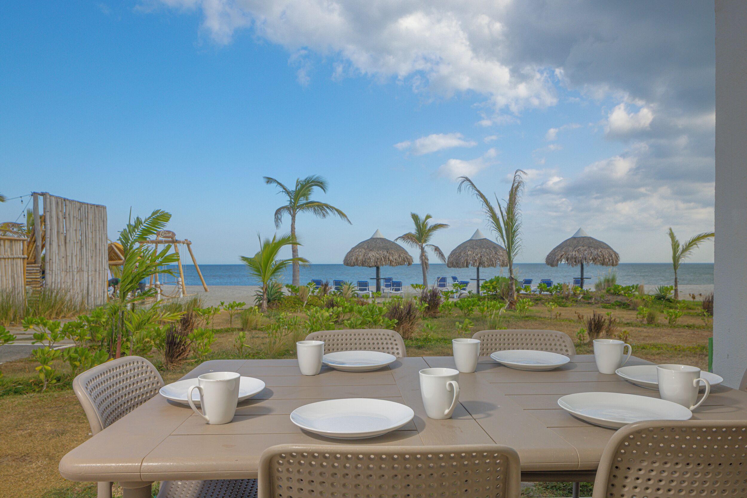 Un espace de restauration en plein air avec une table dressée pour quatre personnes, donnant sur une plage bordée de parasols en chaume et de palmiers.