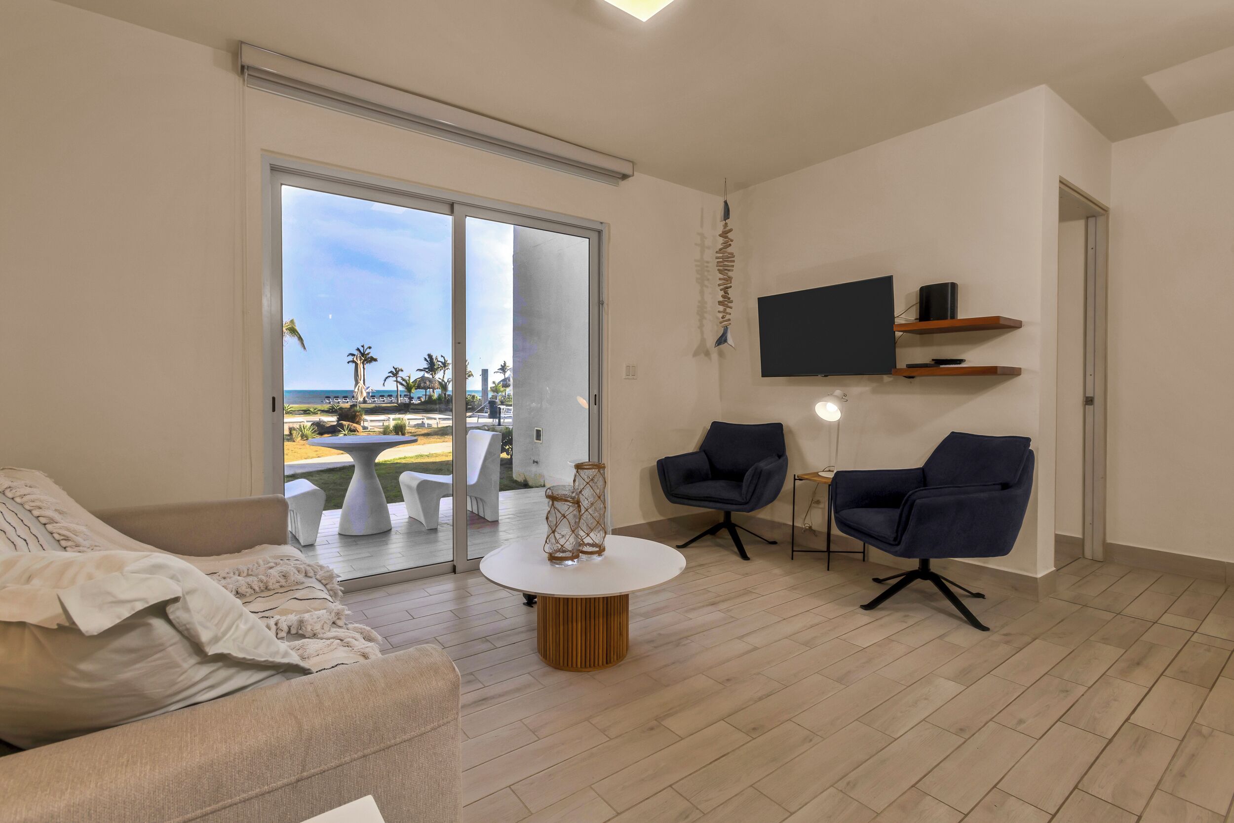 Una sala de estar moderna con un amplio ventanal corredero que da a un balcón, una mesa de centro redonda de madera, dos sillones oscuros y un televisor de pantalla plana montado en la pared.