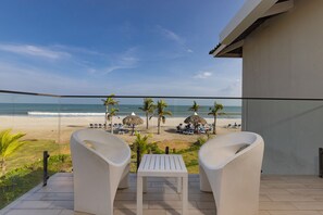 Un balcon avec deux chaises blanches et une petite table, donnant sur une plage bordée de palmiers et ombragée par des parasols en feuilles de palmier.
