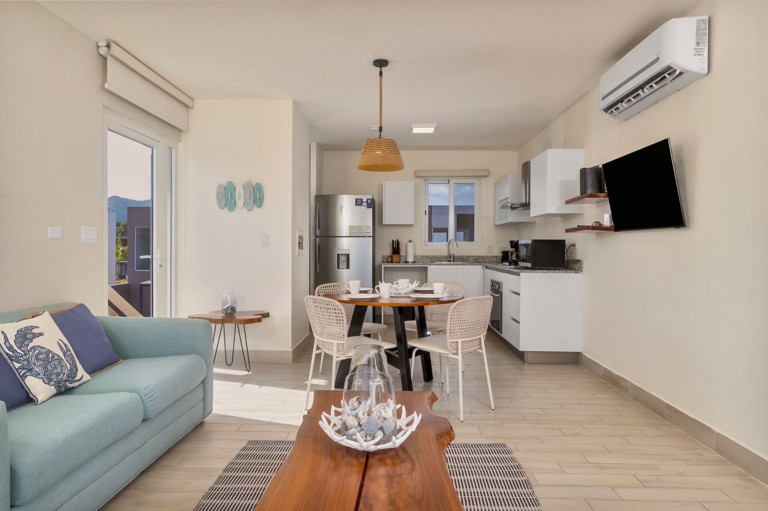 Una sala moderna con sofá, una mesa de centro y un comedor. Hay una cocina con refrigerador, estufa y microondas. Un televisor está montado en la pared, y se ve un aire acondicionado.
