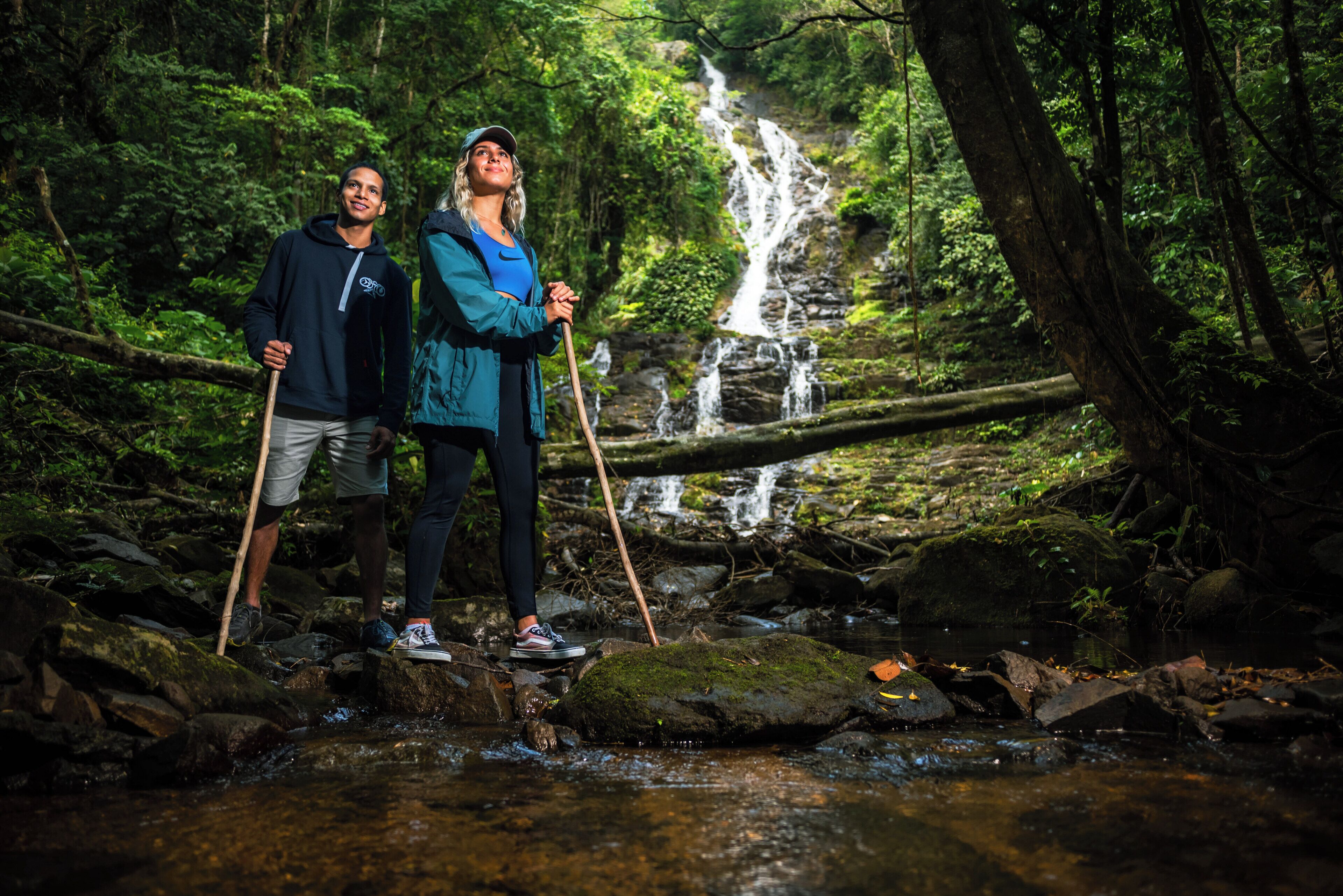 Dos personas de pie sobre rocas junto a una cascada en un bosque.