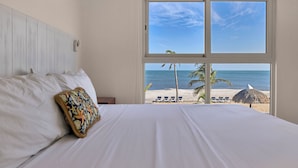 Habitación de hotel con una cama grande, vista a la playa y una sombrilla de paja.