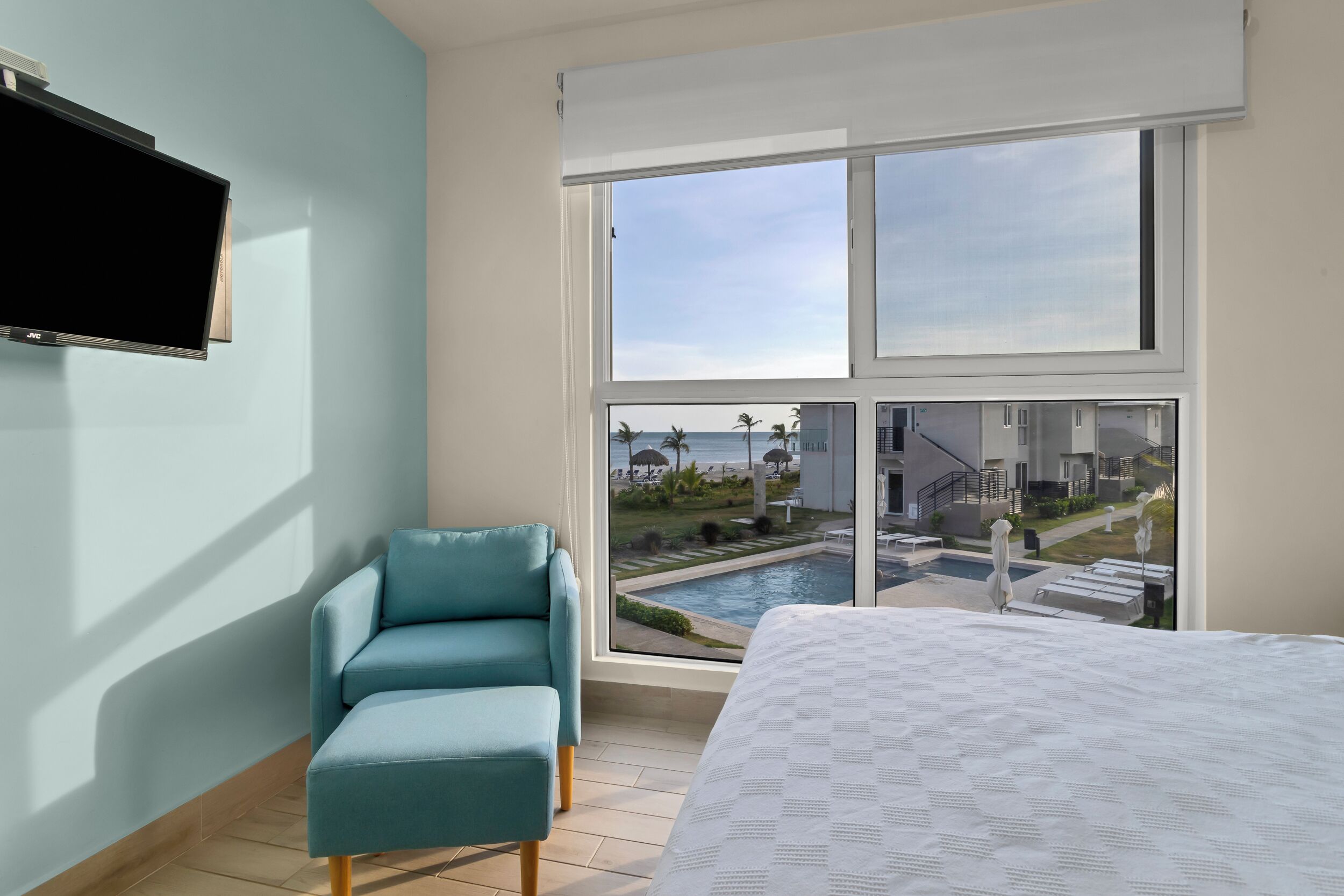 Una habitación de hotel moderna con un ventanal grande, una cama, un sillón azul y un televisor de pantalla plana.