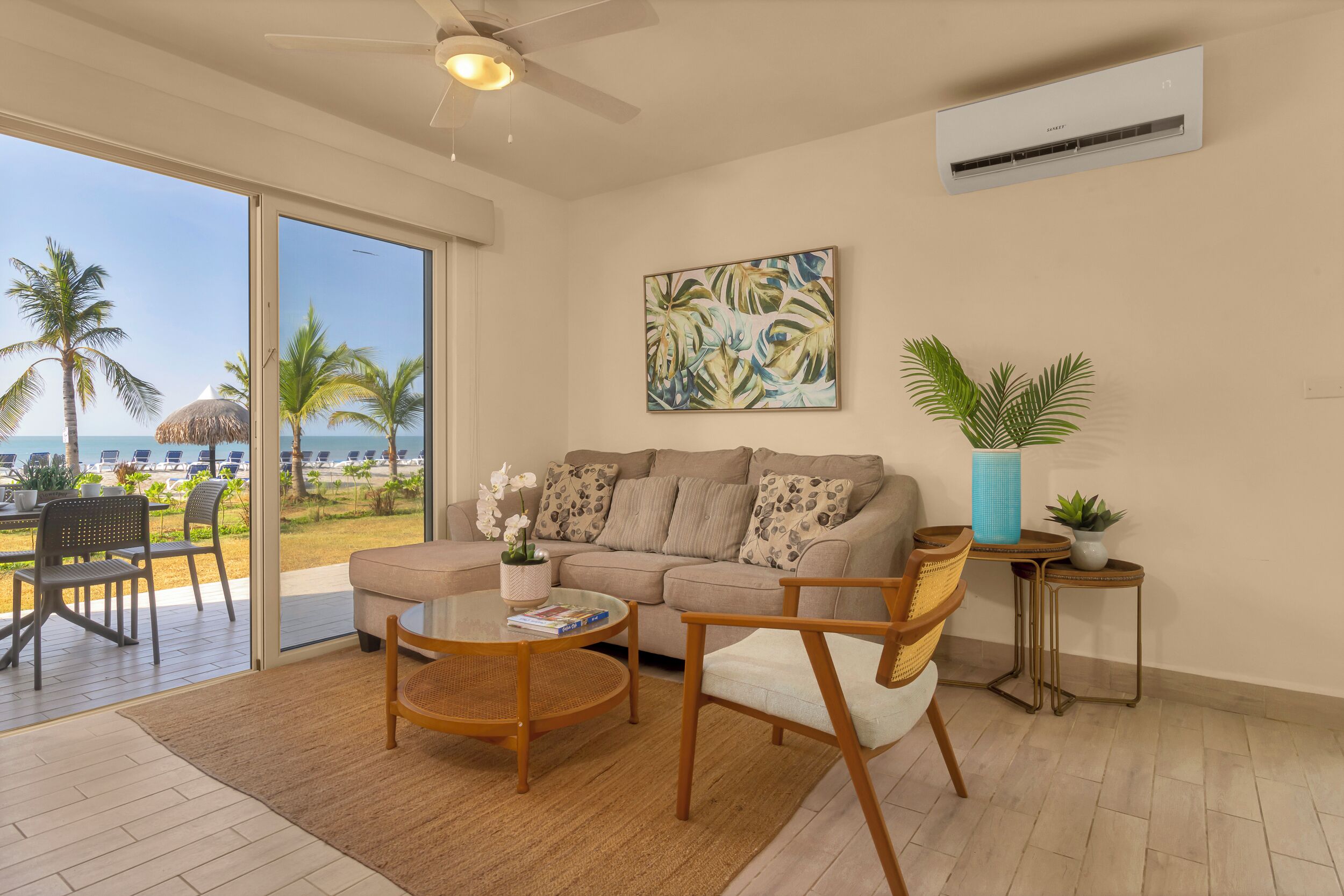 Una sala de estar con sofá, mesa de centro y sillas, un ventanal con vista a la playa y un equipo de aire acondicionado en la pared.