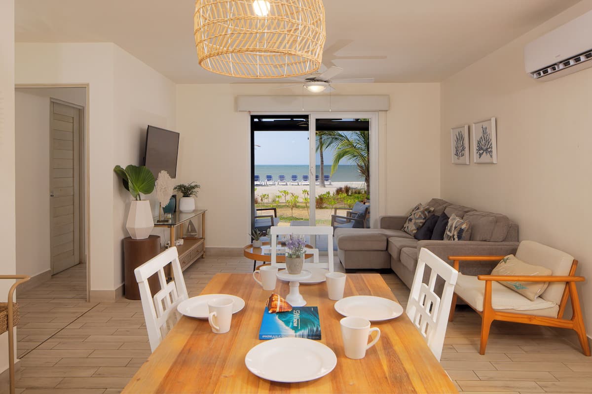 Una sala de estar moderna con una mesa de comedor de madera preparada para cuatro, un sofá y vista a la playa.