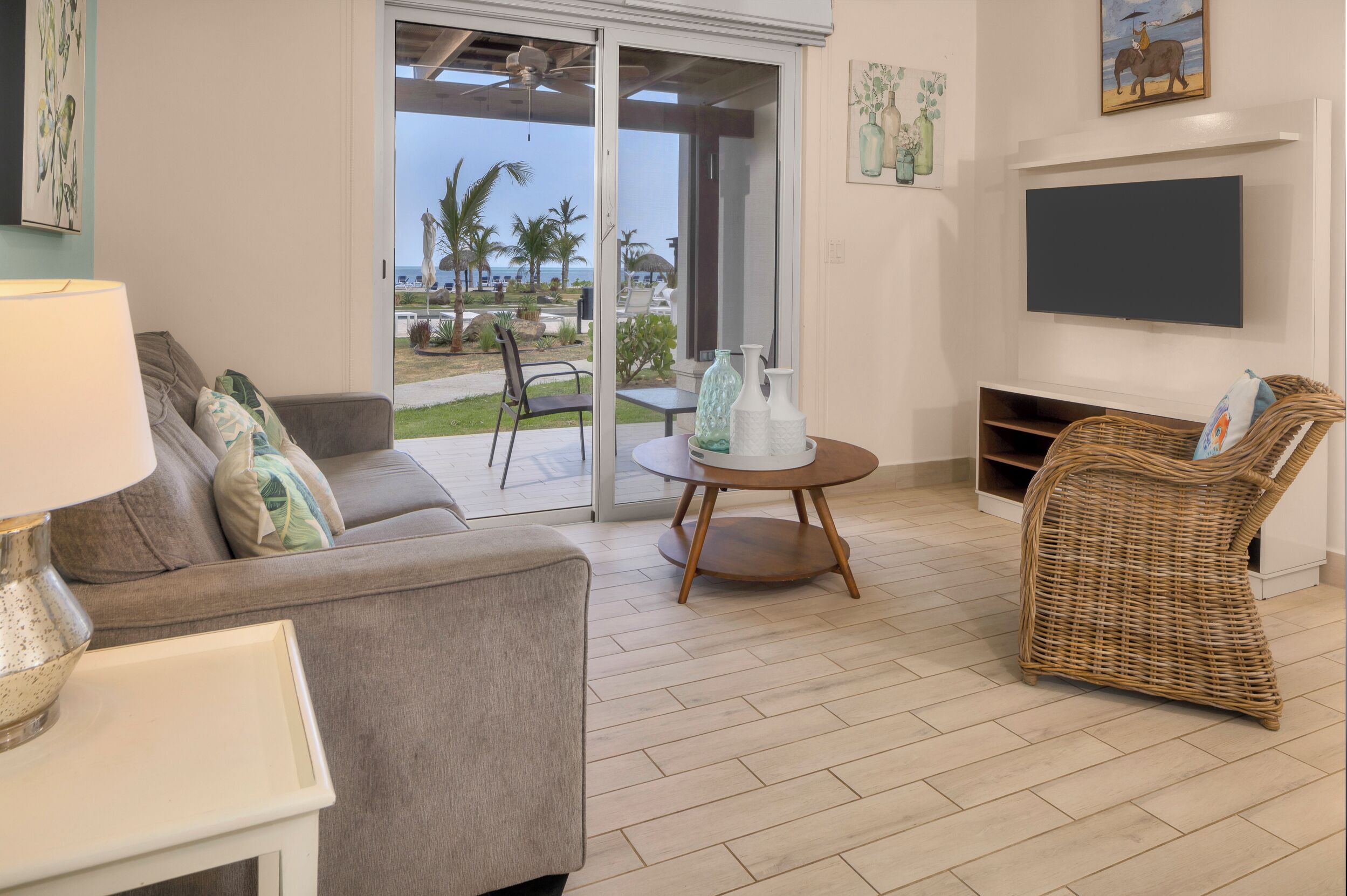 Una sala de estar con un sofá, una mesa redonda con jarrones decorativos, un sillón de mimbre y un televisor montado en la pared.