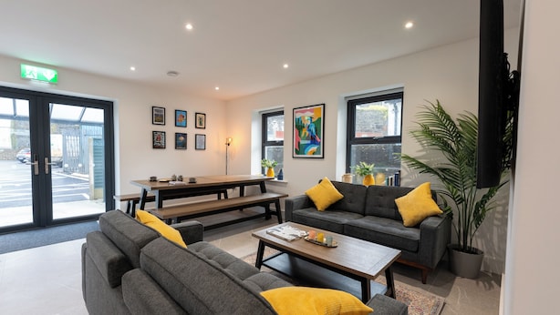 Een moderne woonkamer met grijze banken en gele kussens, een houten salontafel en een bankje. Er hangen kunstwerken in lijsten aan de muren en een groot raam met uitzicht op buiten.