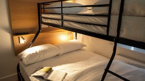 Een stapelbed met een houten hoofdbord, een nachtkastje met een boek, en een wandlamp.