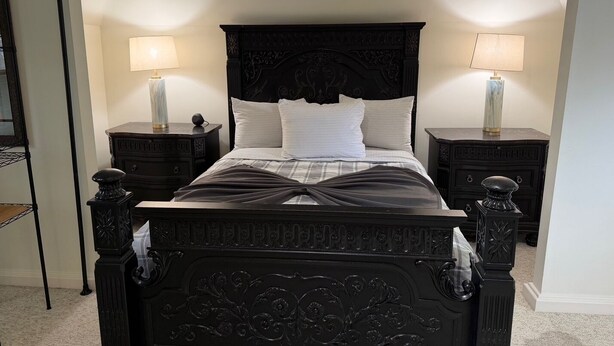 Un dormitorio con una cama negra ornamentada, dos mesitas de noche con lámparas y un espejo grande en la pared izquierda.