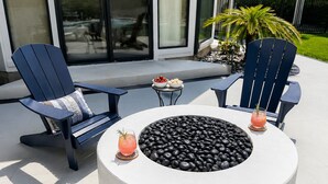 Un espacio exterior moderno con dos sillas Adirondack, una fogata redonda y dos bebidas sobre posavasos.