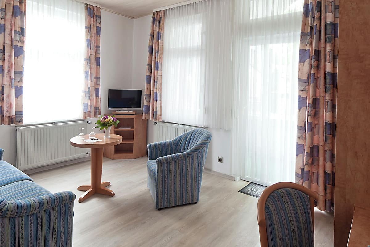 Ein Hotelzimmer mit zwei blau gestreiften Sesseln, einem Holztisch mit Blumenvase, einem Fernseher auf einem Holzschrank und großen Fenstern mit gemusterten Vorhängen.