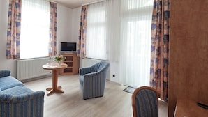 A hotel room with two blue-striped armchairs, a wooden side table with a vase of flowers, a television on a wooden stand, and large windows with patterned curtains. - Hotel Flora (Bad Harzburg)