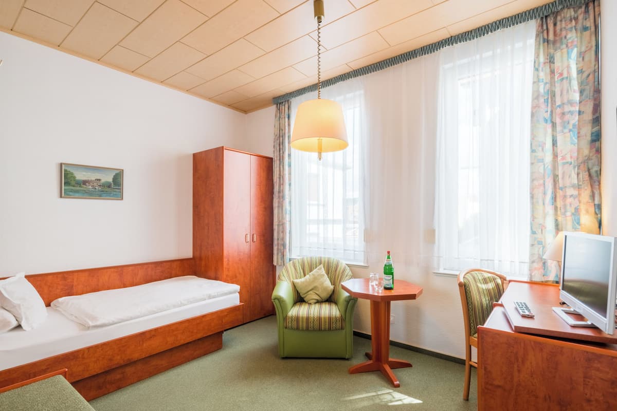 Ein Hotelzimmer mit einem Einzelbett, einem Holzschrank, einem grünen Sessel, einem kleinen Tisch mit einer Flasche, einem Stuhl und einem Schreibtisch mit einem Computer.