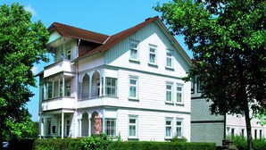 A large, two-story building with a white exterior and a brown roof, surrounded by lush green trees. - Hotel Flora (Bad Harzburg)