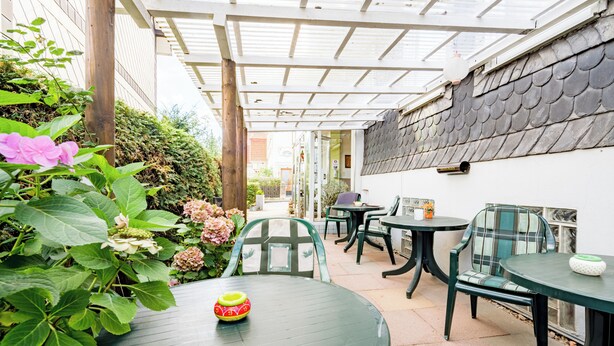 A covered patio area with tables and chairs, surrounded by plants and a decorative lantern. - Hotel Flora (Bad Harzburg)