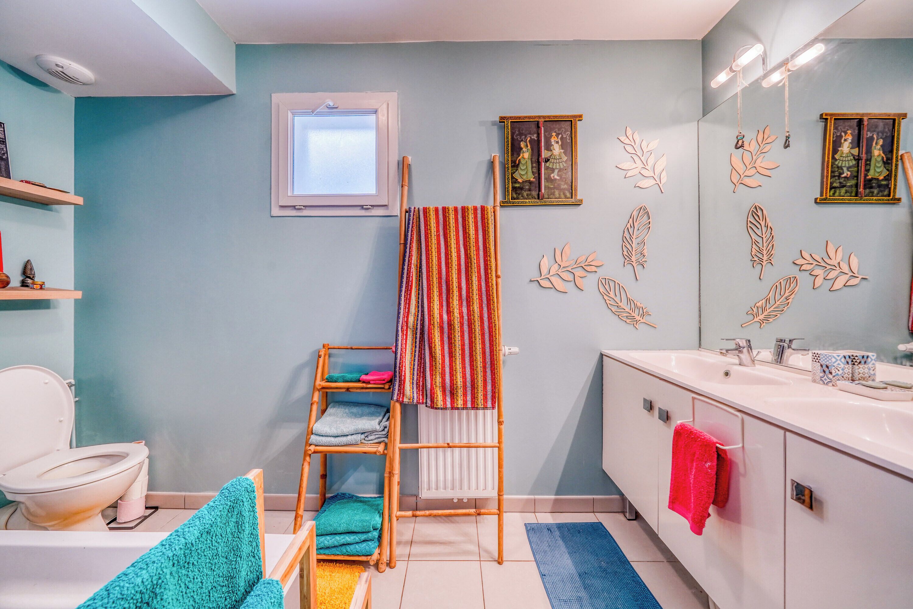 Une salle de bain avec un lavabo blanc, un miroir, une fenêtre et une étagère contenant des articles de toilette.