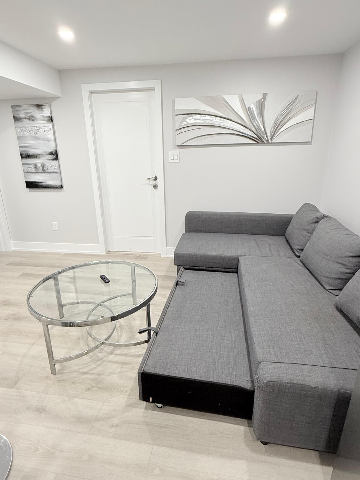 Een moderne woonkamer met een grijze hoekbank, een glazen salontafel, en een deur met daarboven een abstract schilderij.