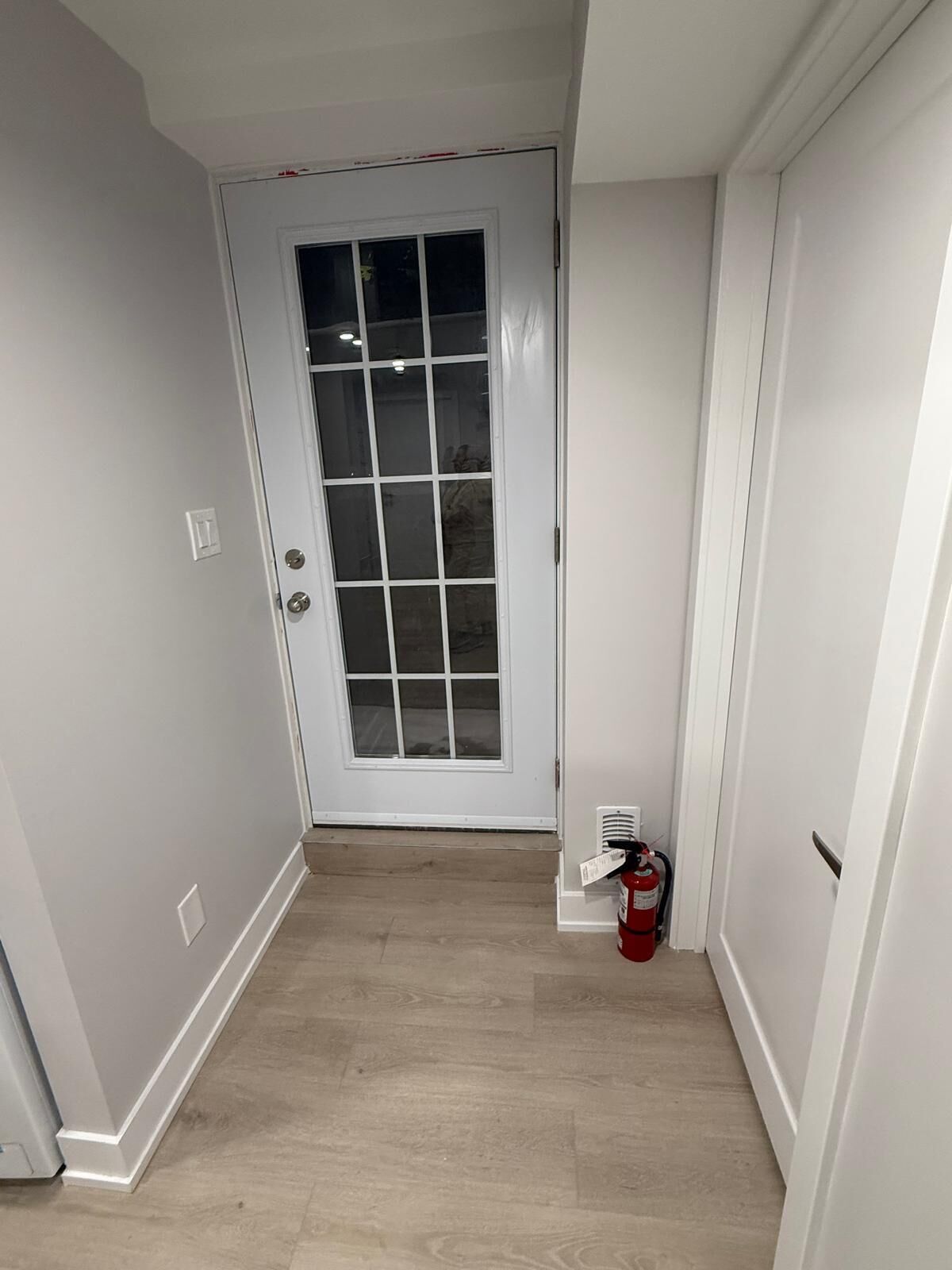A narrow hallway with a white door, light wooden flooring, and a fire extinguisher near the door.