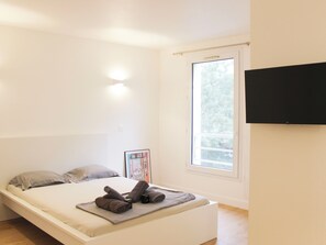 Une chambre à coucher au style minimaliste, avec un lit, une fenêtre et un téléviseur à écran plat fixé au mur.