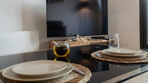 Un coin repas moderne avec une table noire, deux assiettes, un verre et une bouteille d’huile d’olive. Un téléviseur à écran plat est fixé au mur derrière la table.