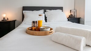 Un lit bien fait, avec du linge de lit blanc, un plateau en bois contenant des croissants et une bouteille de jus d’orange, et un bol de céréales.