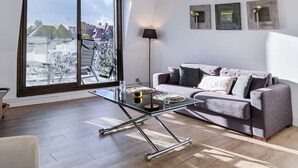 Un salon moderne avec une table basse en verre, un canapé gris et une vue sur les immeubles à travers de grandes fenêtres.