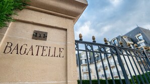 Un bâtiment portant l’inscription « BAGATELLE » sur un mur de couleur beige, une grille en métal ornée de motifs décoratifs et un style architectural classique agrémenté d’un balcon.