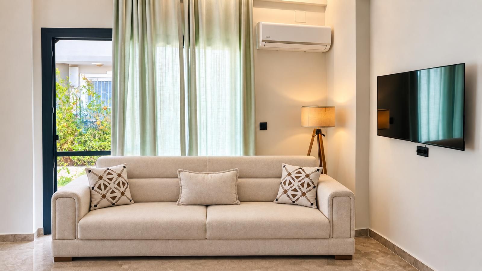 A neutral-toned sofa with patterned cushions, a floor lamp, and a flat-screen TV mounted on the wall.