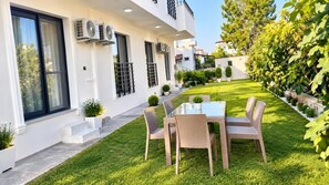 Un área de estar moderna al aire libre con una mesa de vidrio y sillas de mimbre, rodeada por un césped cuidado y plantas en macetas.
