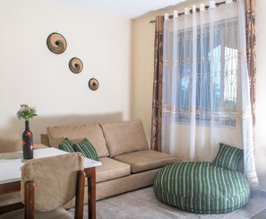 Ein Wohnzimmer mit einem beigen Sofa, einem gemusterten Hocker, einem kleinen Tisch mit einer Vase und dekorativen Wandgestaltungen.