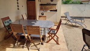 Eine Terrasse im Freien mit einem runden Tisch, Holzstühlen, einem eingebauten Grill und einer Lounge-Ecke mit Liegestühlen.