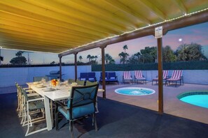 Een terras buiten met een overdekte eethoek, zwembad en lounge stoelen.