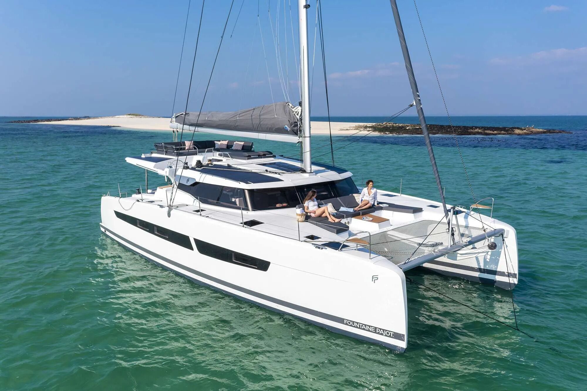 A white catamaran with two people on deck, a covered seating area, and a view of a beach and rocky outcrop in the background.
