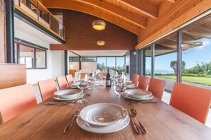 A dining area with a long wooden table set for a meal, large windows offering a view of the outdoors, and a high ceiling with wooden beams. - Casa Vista Azul by Atlantic Holiday (Santa Cruz)