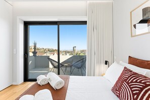A hotel room with a large bed, a view of the city through a sliding glass door, a small balcony with a chair and table, and a window with curtains. - GuestReady - Porto Retreat with Terrace (Porto)