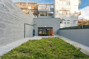 En moderne bygning med en betonfacade, en glasdør og et lille haveområde med græs og planter.