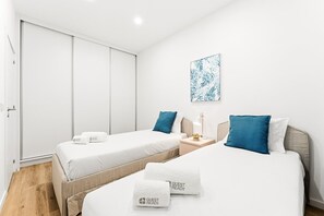 Et hotelværelse med to senge, hver med et håndklæde der står 'GUEST READY', et natbord med en lampe, og et kunstværk på væggen.