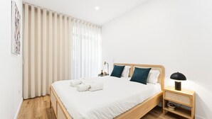 A neatly made bed with white linens and two dark pillows, a bedside table with a lamp, and a window with sheer curtains.