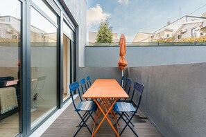En hyggelig udendørs terrasse med et træbord og blå og orange stole, en orange parasol samt en glasdør, der fører ind i et rum.