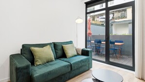 A green sofa with cushions, a small round table, and a sliding glass door leading to an outdoor seating area with blue chairs and an orange table.