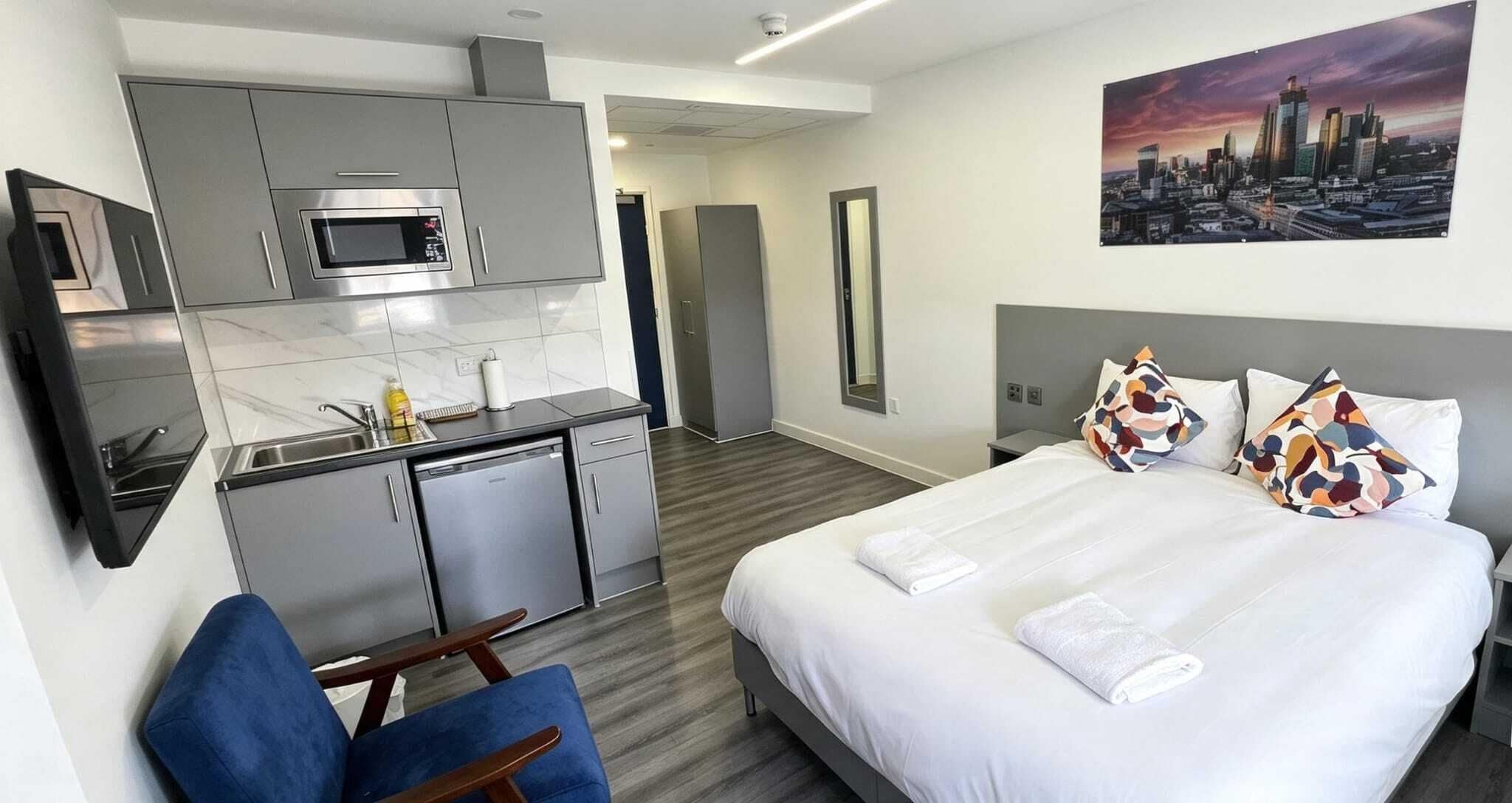 A modern hotel room with a bed, a desk, a microwave, and a cityscape painting.