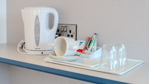 Una kitchenette con cafetera, una taza, un plato y un recipiente de vidrio con utensilios.