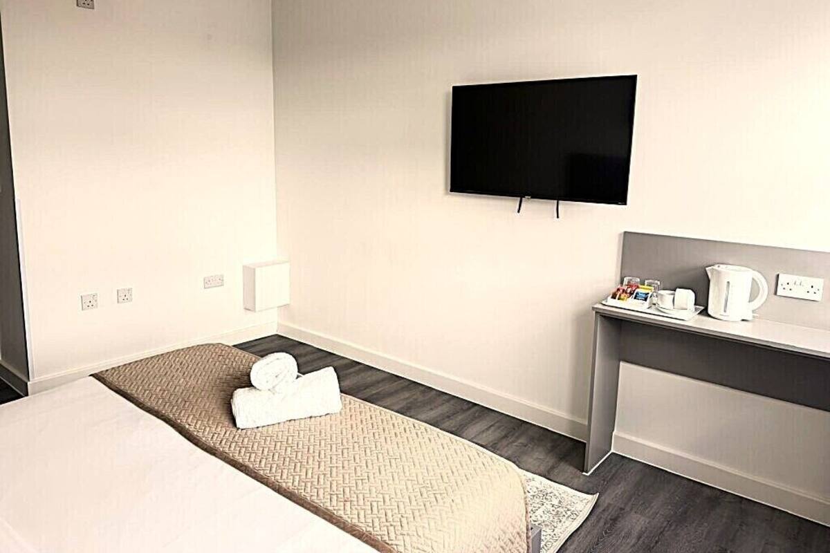 Quarto de hotel com cama, televisão de tela plana, pequena mesa de trabalho com chaleira e mesa de cabeceira.