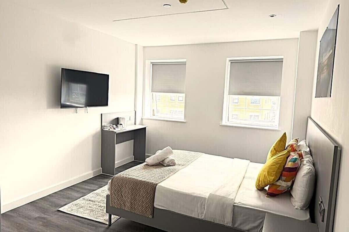 Quarto moderno com cama, mesa de cabeceira, televisão e janelas.