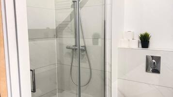 A modern bathroom with a glass-enclosed shower, a toilet, and a small shelf with toiletries.