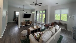 Amplia sala de estar con un sofá grande, una mesa de centro y un televisor montado en la pared. Hay dos ventiladores de techo, un ventanal con persianas y una puerta que conduce a otra habitación.